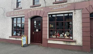 Brown and cream exterior of CF Beattie with sign stating Shooting & Fishing Accessories / Gunsmith plus antique displays in windows