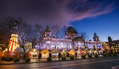 Belfast City Hall light up at night with Christmas market stalls in foreground in front of the City hall front gates.  
