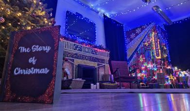 a quiet living room scene is depicted lit up with Christmas lights and a tree. a book is propped up on the floor and the front cover reads 'The Story