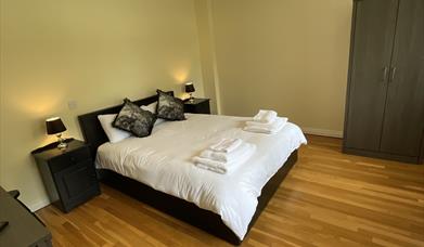 Double bedroom with hard floor wooden floor. Double bed dressed in white linen, bedside cabinets on both sides of the bed. Wardrobe and side of dressi