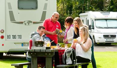 Image shows family and friends sitting at picnic table with caravan in the background
