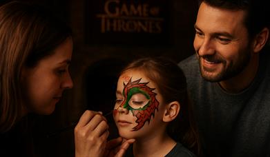 Woman painting a dragon on child's face as father watches.