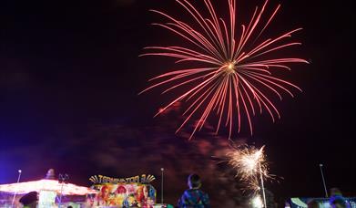 Image shows fireworks lighting up the night sky with the crowd and fairground amusements below