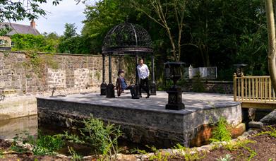 A man and woman resting under a canopy on a small manmade island in a garden.