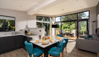 Open plan kitchen and dining area with view of lounge and outside area in background. Open patio doors floor to ceiling leading onto decked patio area