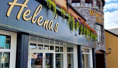 Exterior of Helena's Coffee Shop and Bistro, featuring a modern black and white facade with gold lettering. Colorful hanging plants decorate the top o