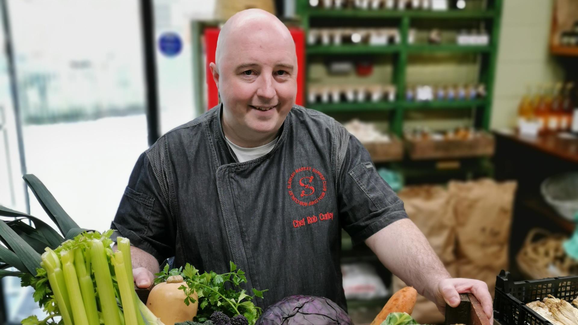 a man stands with a box filled with fresh vegetables including carrots, parsnips, celery, onions and cabbage