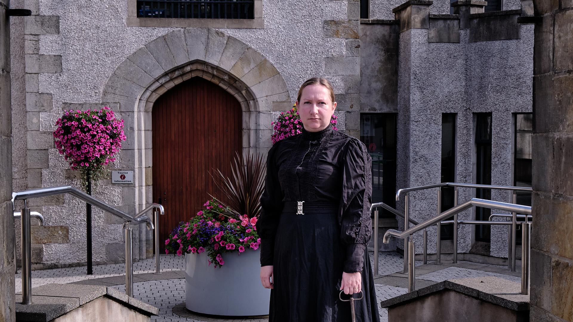 Image of woman standing under the stone arch of the Bridewell, Magherafelt.  She is dressed in a long black dress from 1800's and holds a large brass