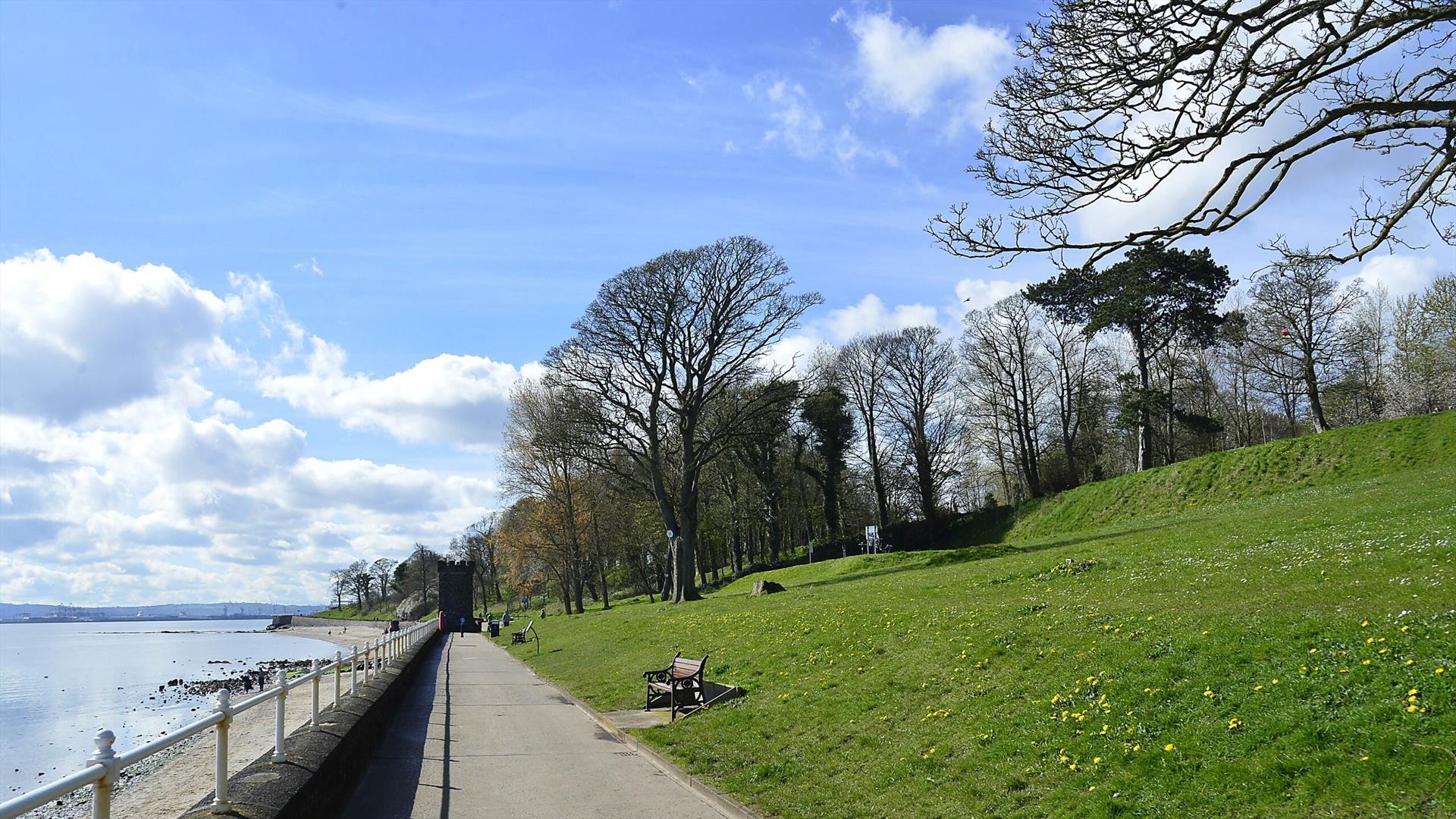 A paved pathway alongside the Belfast Lough shoreline in Newtownabbey, with green grass, benches, and the lough stretching into the distance.