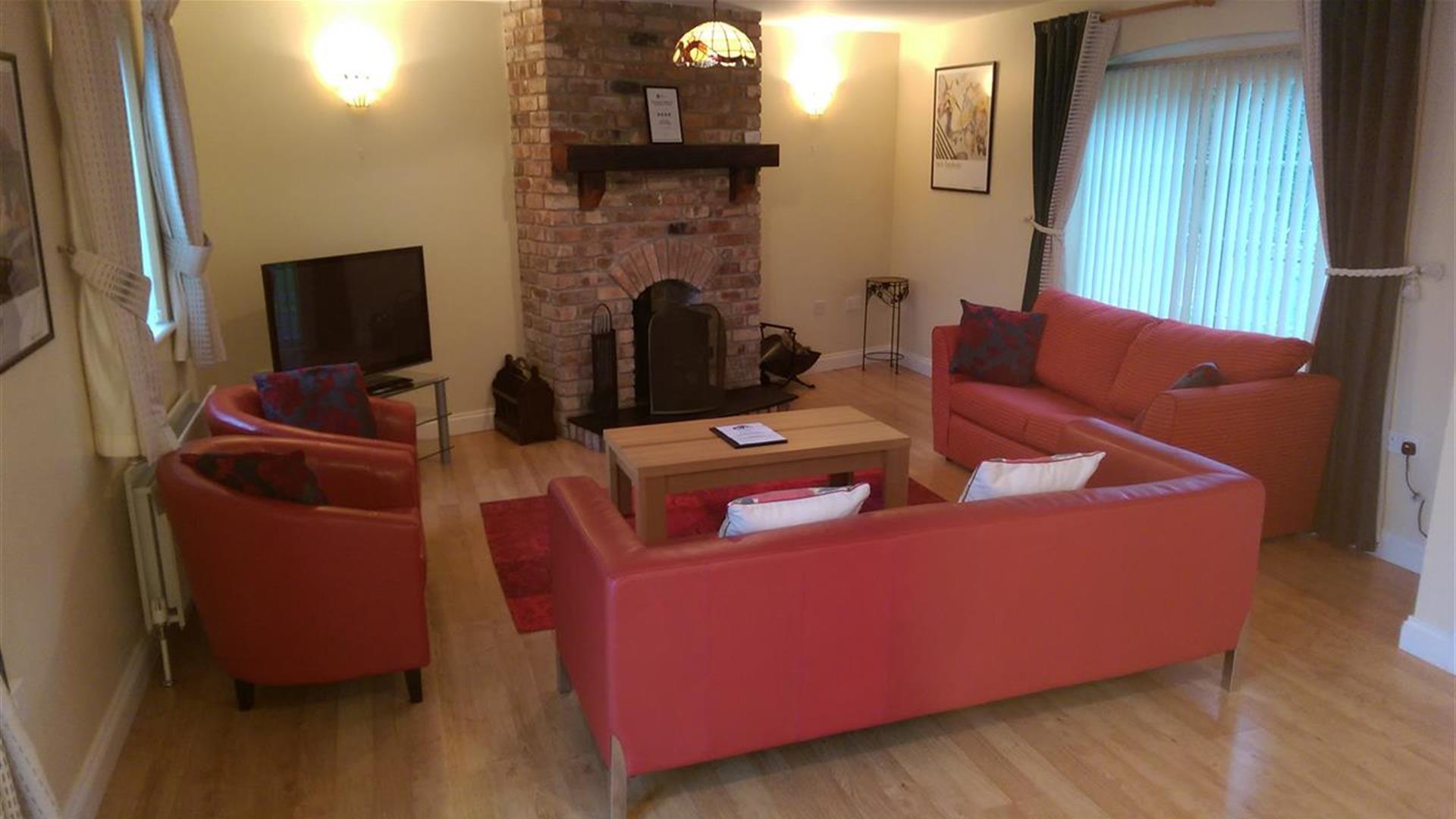Image shows lounge area with sofas, chairs, TV and fireplace. Two windows in the room.