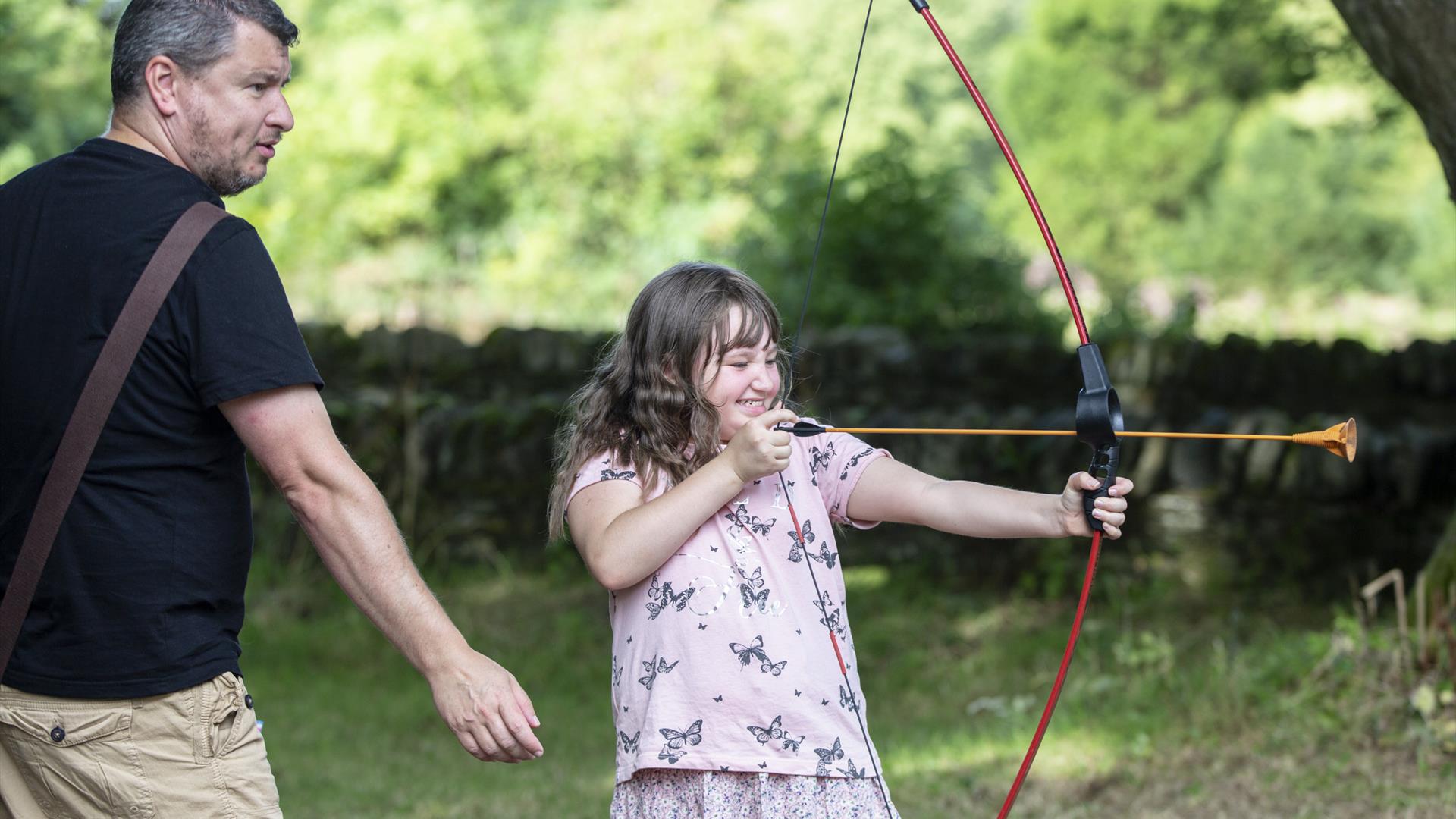 Girl shooting bow and arrow with her father behind her.