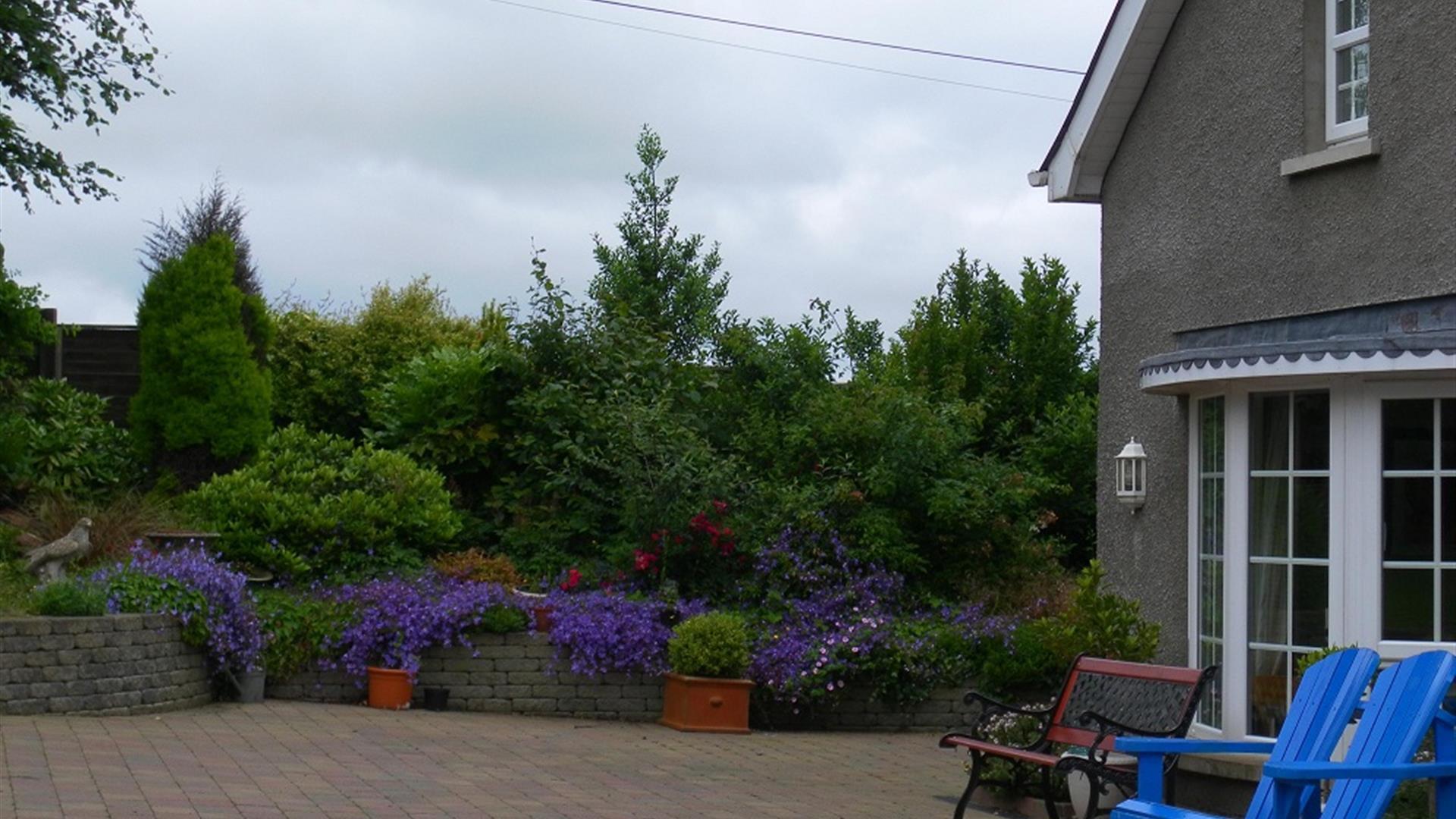 A paved area with garden furniture, trees and purple flower bushes.