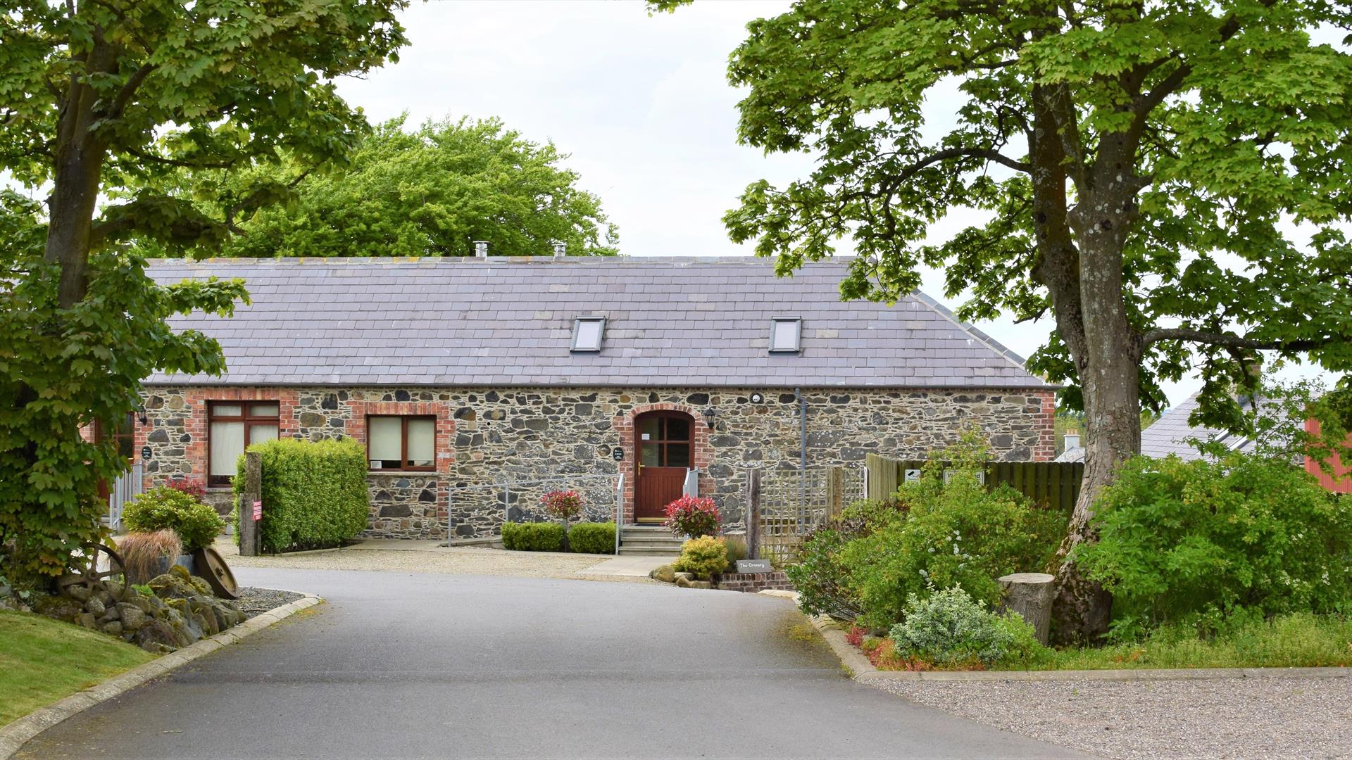 Image shows front of the property. Tarmac driveway bordered by trees on both sides. There is a ramp up to front door and also a path with some low ste