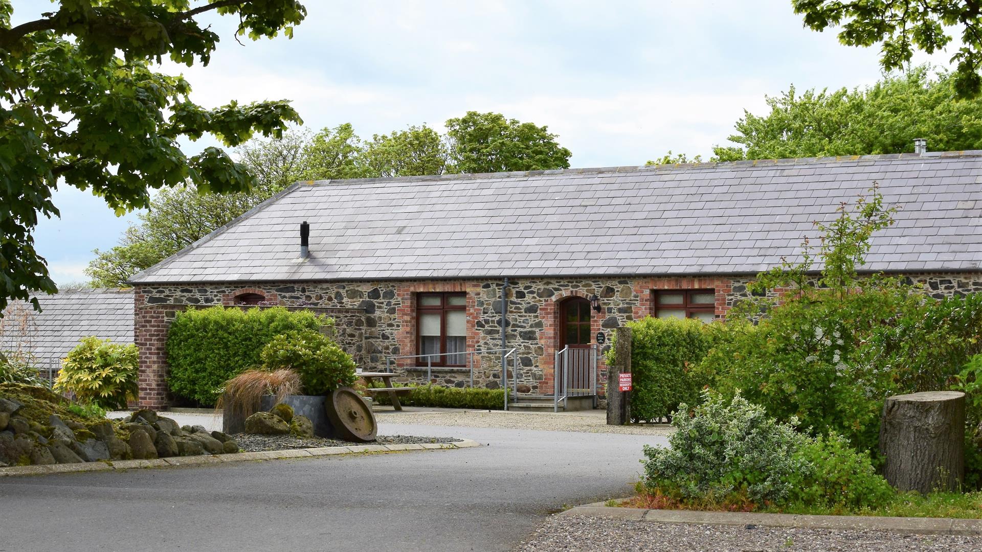 Image shows outside of stone built property with wooden picnic table in small courtyard. Hedges and trees in foreground.