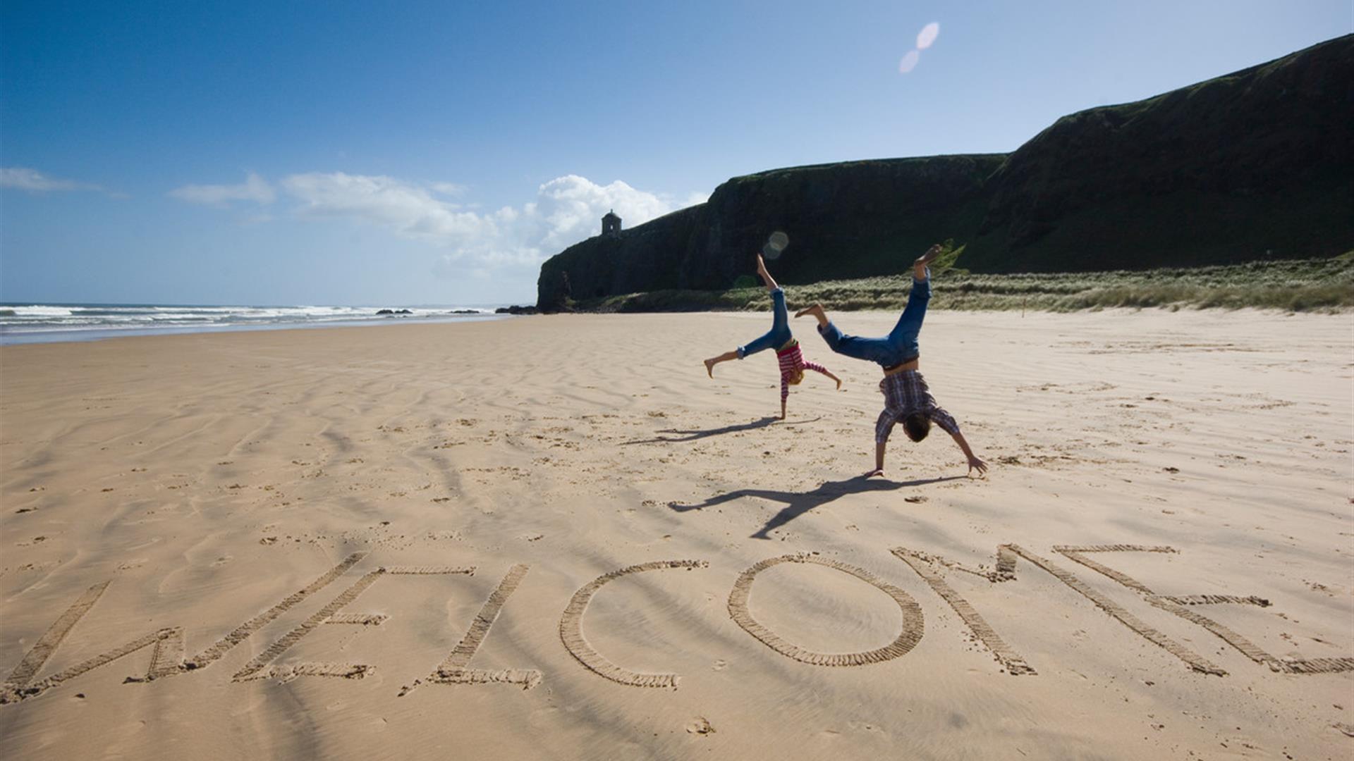 Image of beach with cliffs and sea in the background. The iconic Mussenden Temple is visible in the distance. In the foreground, 'welcome' has been ca