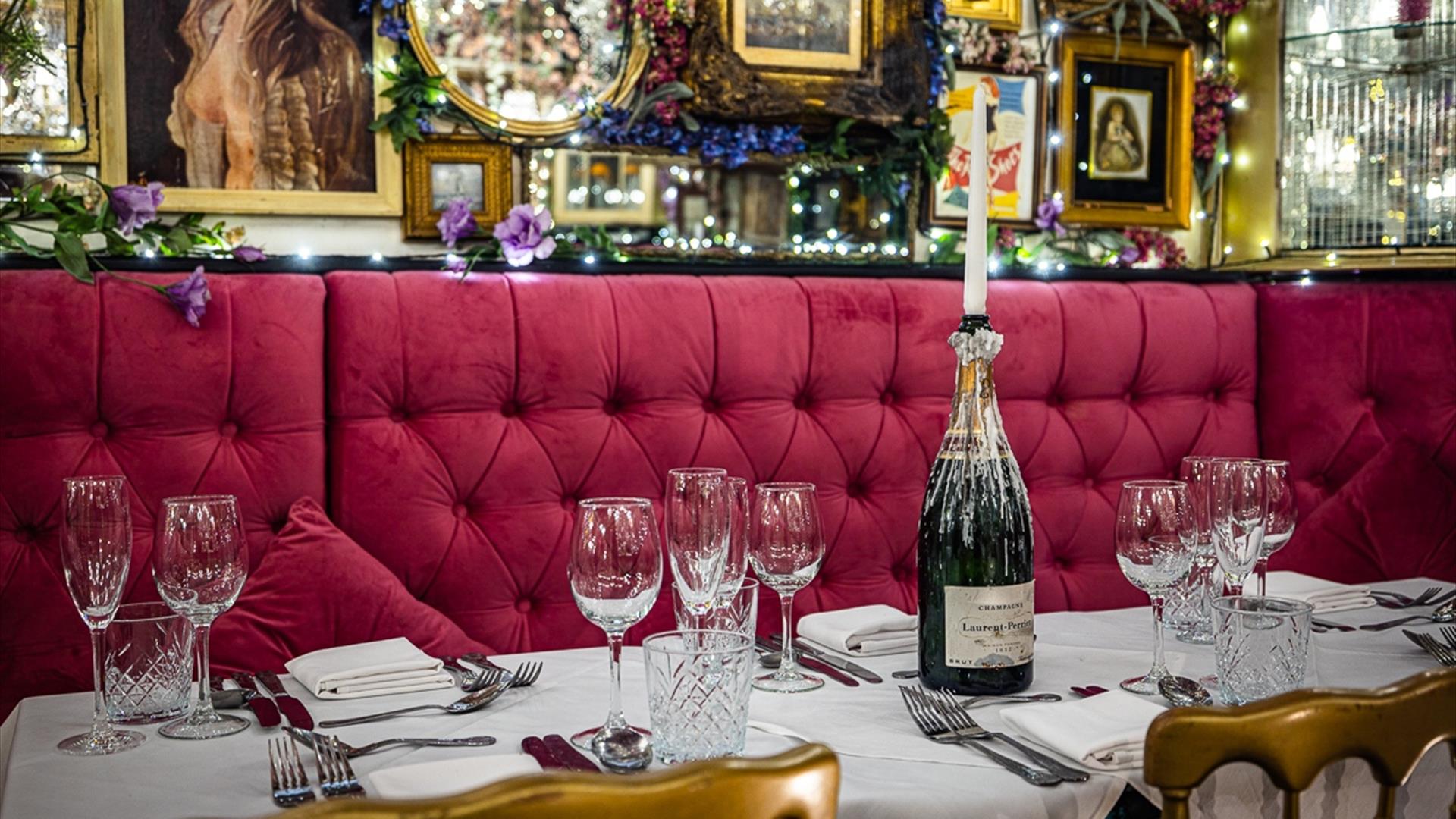 A table with red felt booth seating set with wine glasses and a champagne bottle holding a candle.