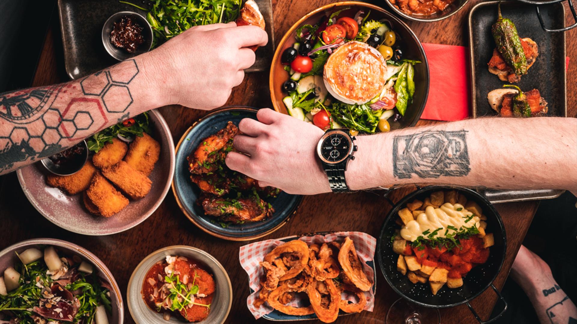 People with tattooed arms reaching over a table for tapas food.