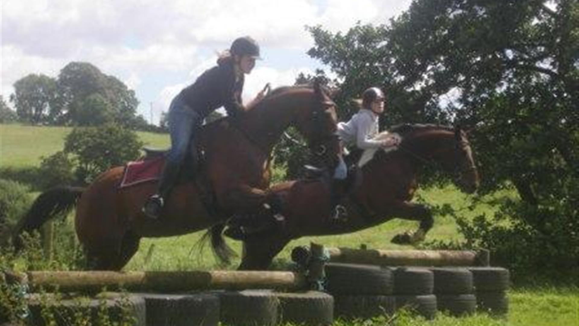 Two horse riders jumping over a tyre and log obstacle.