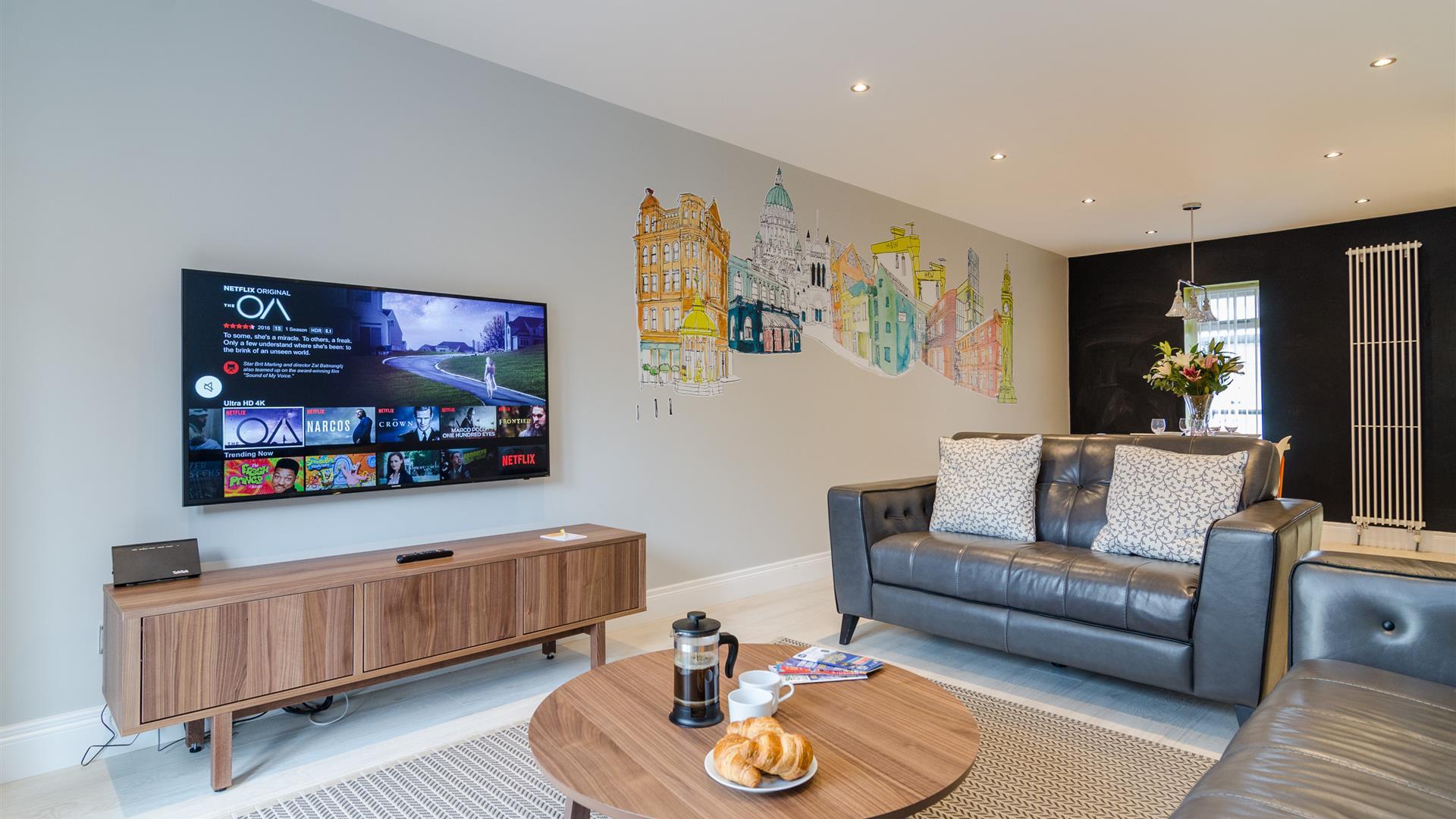 A living room with grey leather sofas, a coffee table with coffee and croissants and a TV with Netflix.