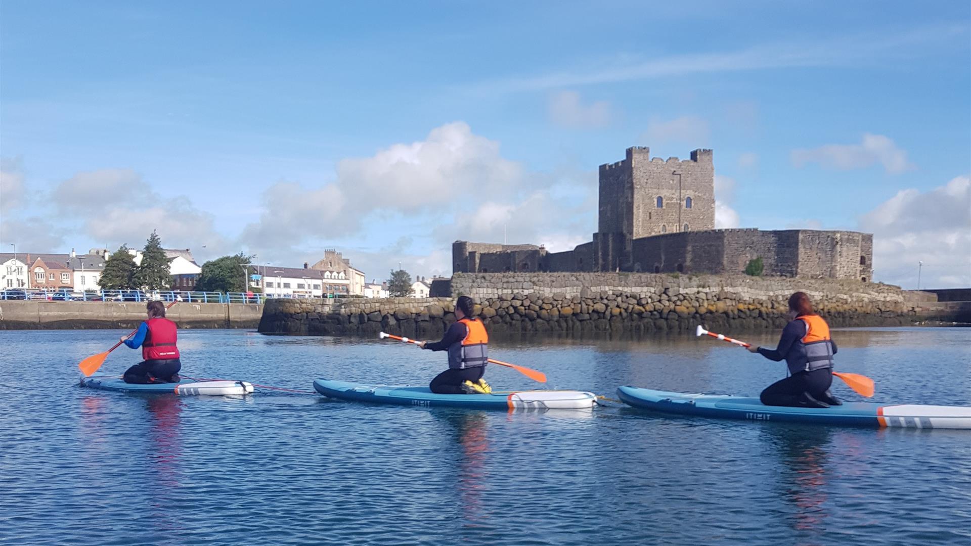 image of 3  paddleboarders kneeling on boards in water in front of Carrickfergus Castle.