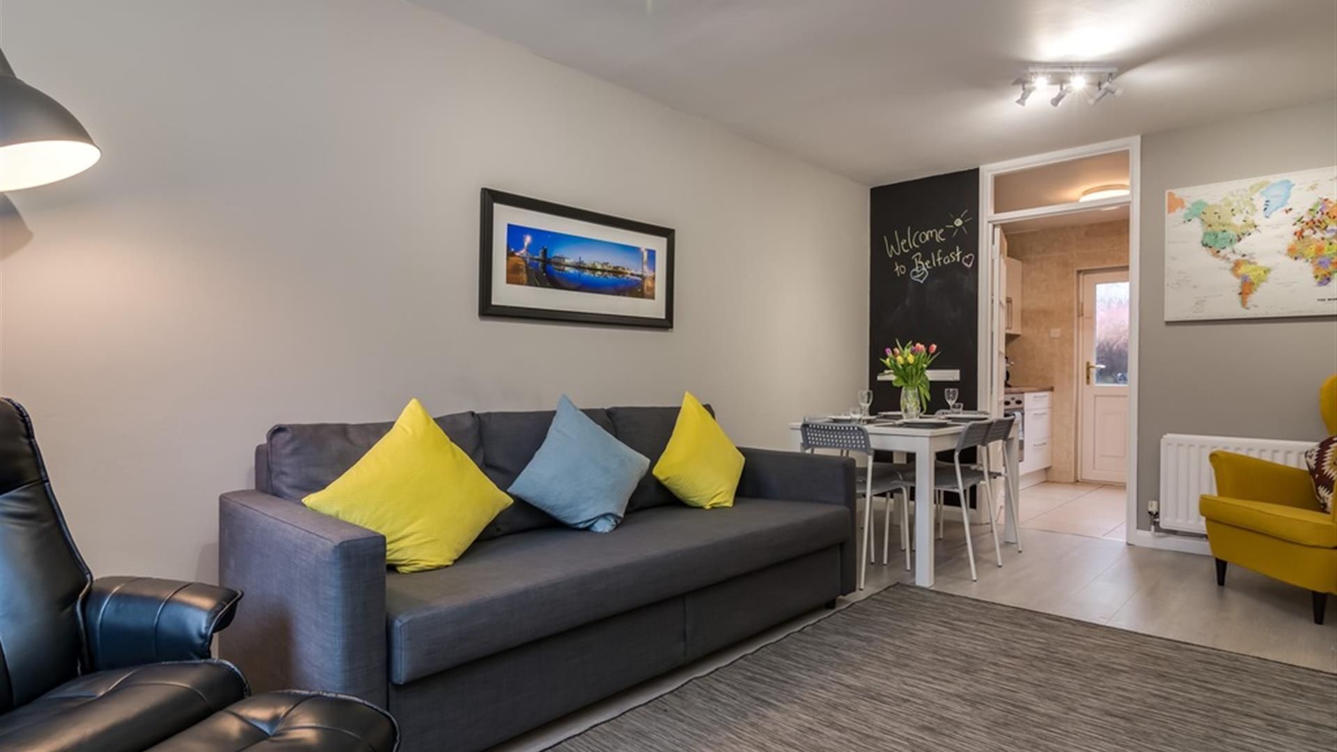 A photo of a living room with a sofa and chair which leads to a dining table and kitchen entrance.