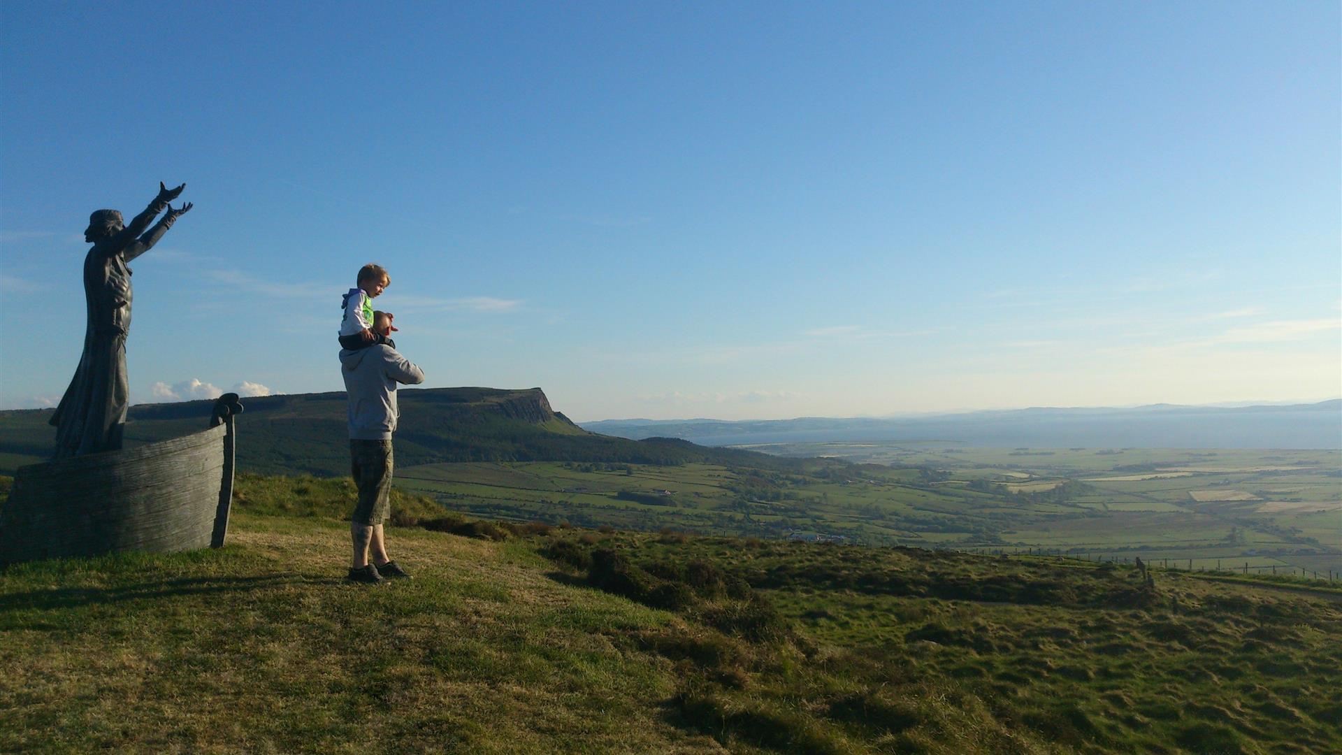 Person standing beside Manannan Mac Lir statue looking at fields and blue sky.