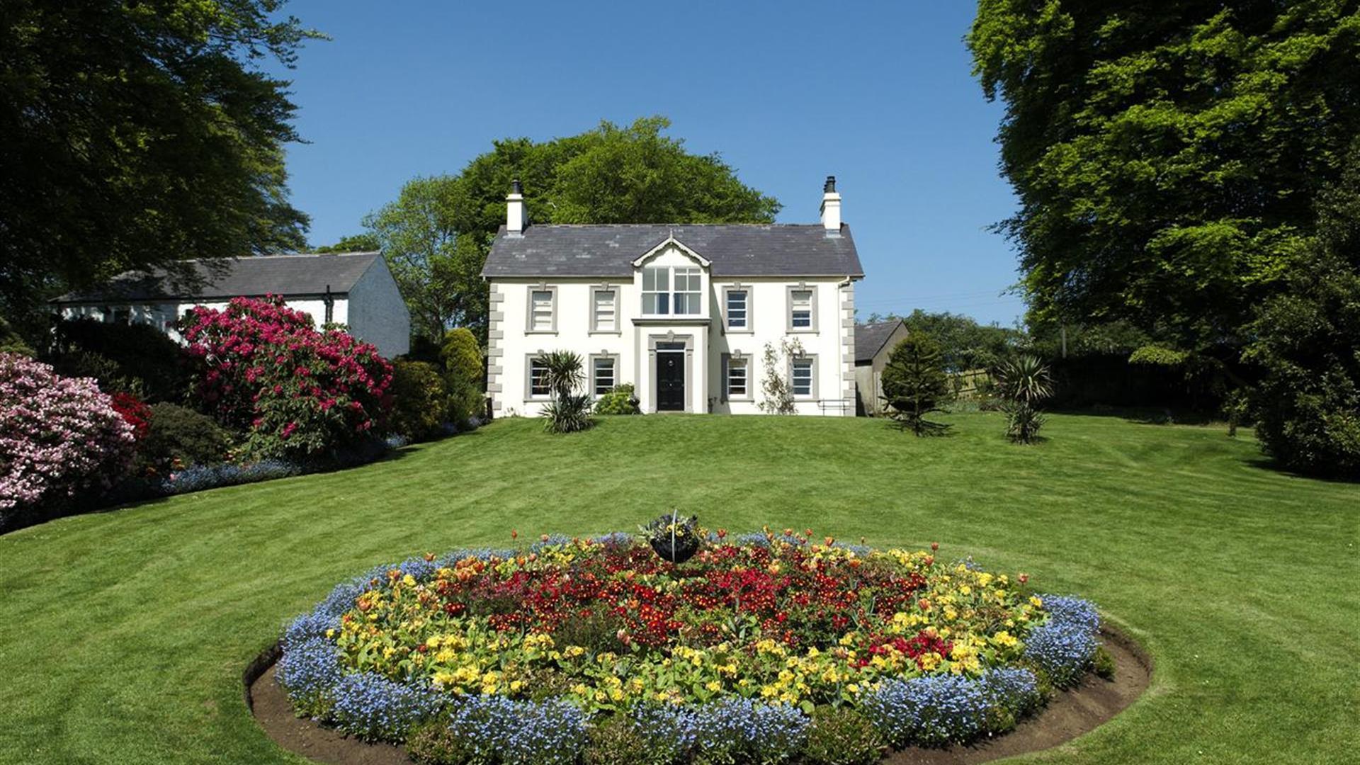 A house at the end of a lawn with a large circular flowerbed.