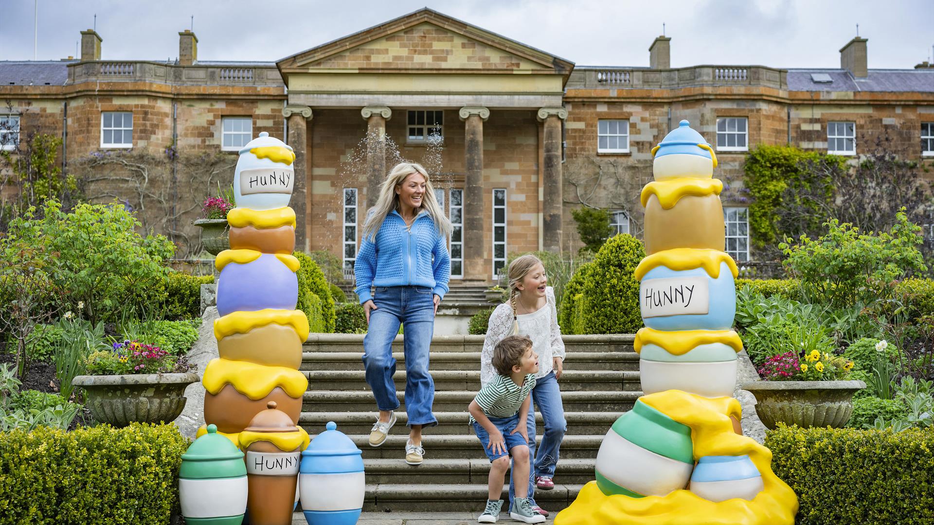 A woman and two children are running down the steps of the South Terrace at Hillsborough Castle. On either side of the steps are brightly coloured sta