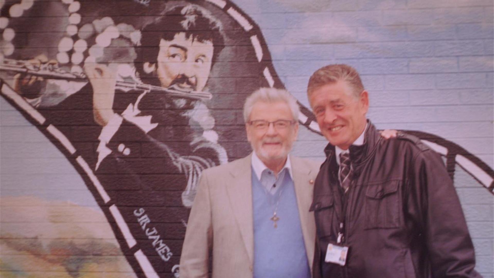 Two men standing in front of a mural of Sir James Galway.