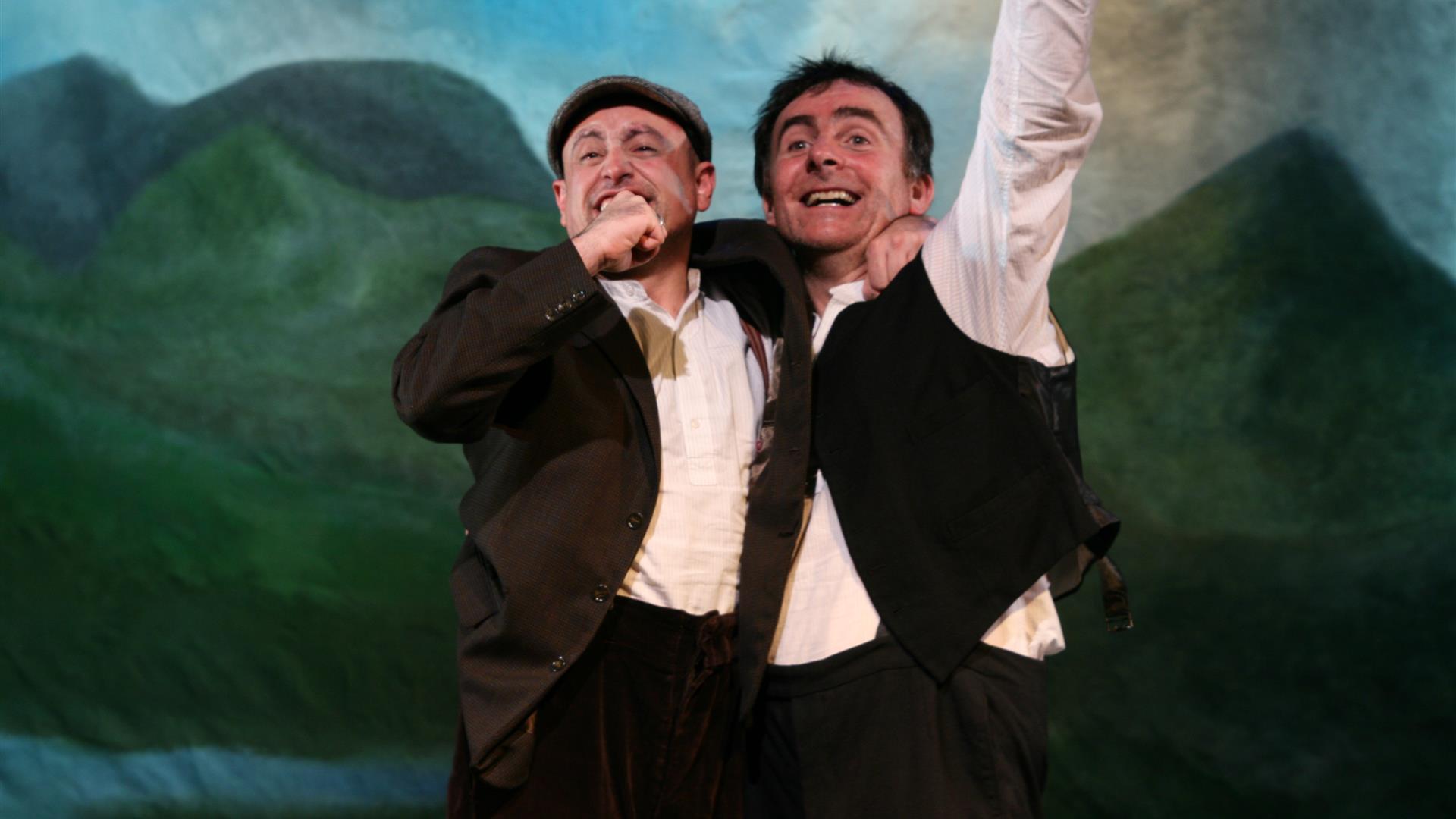 Two joyful men performing on stage in front of a background with hills.