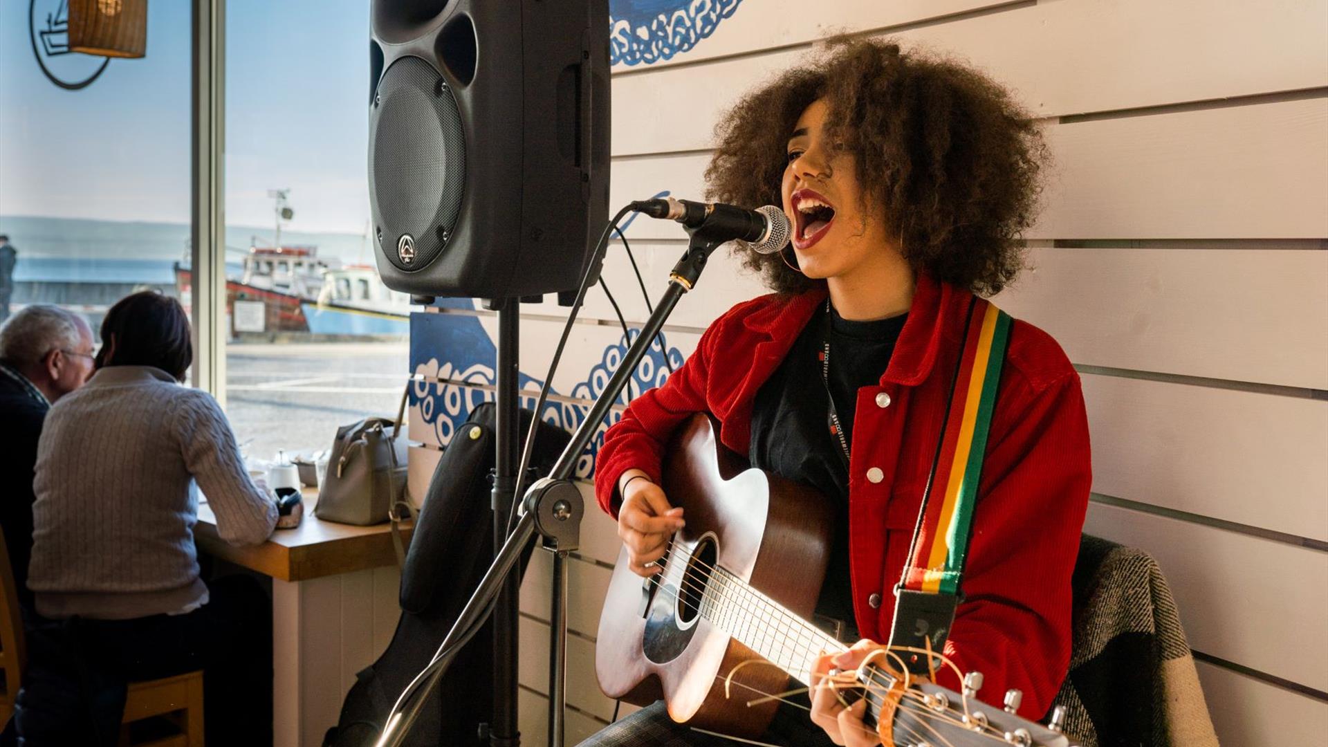 Image shows a lady singing and playing guitar.  In the background there are two people sitting looking out the window at the harbour in Portstewart.