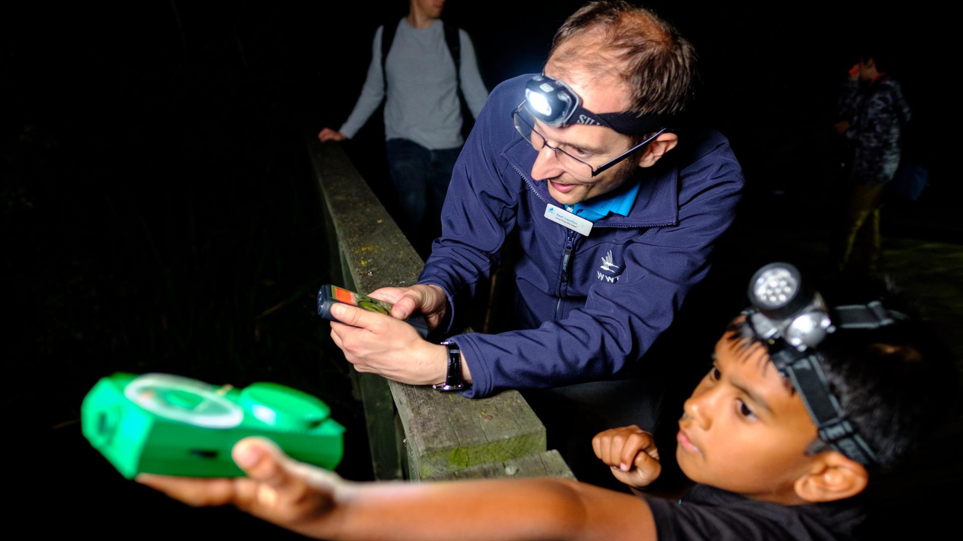 Child wearing a head torch and holding out a bat detector, guided by a member of staff.