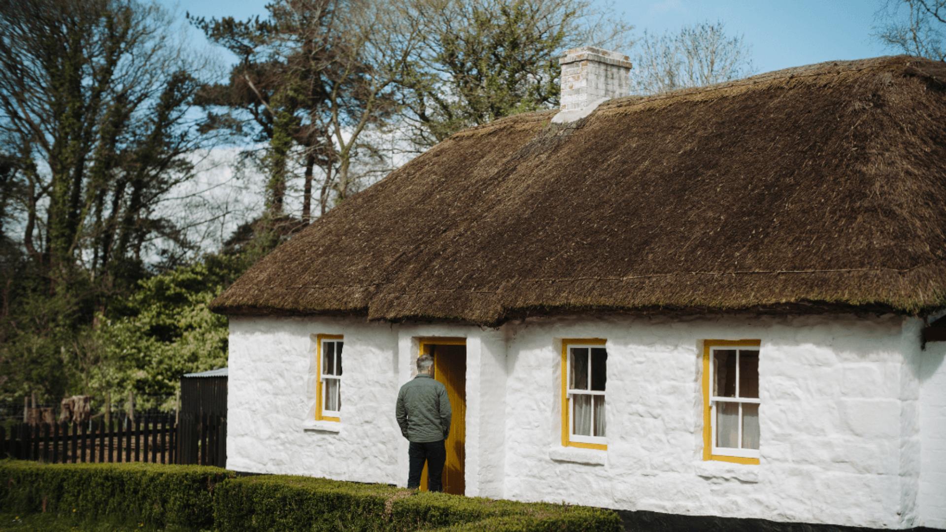 A person standing at the entrance of a white stone cottage with yellow rimmed windows and a thatch roof