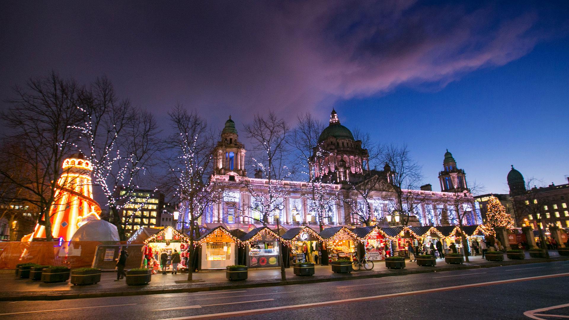 Belfast City Hall light up at night with Christmas market stalls in foreground in front of the City hall front gates.