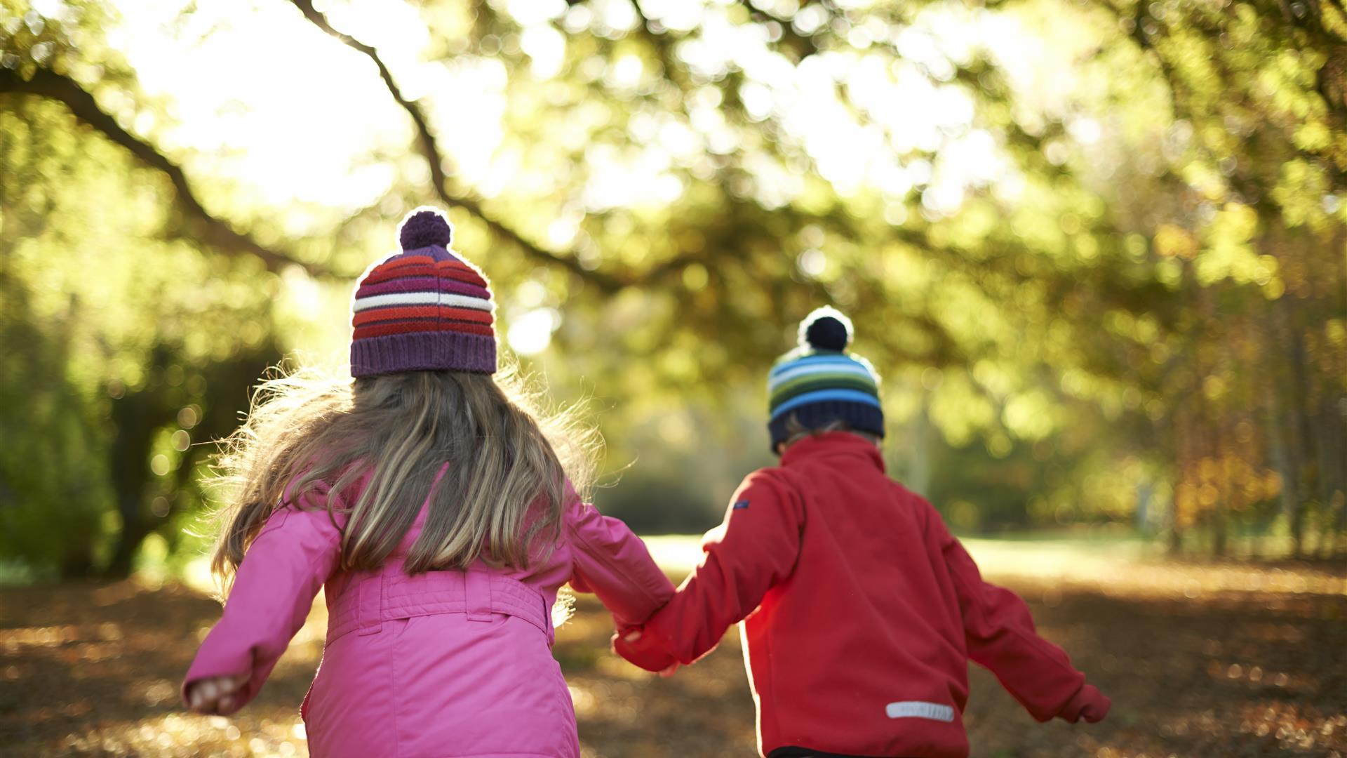 Photo of two children with woolly hats on running through the autumn leaves of the park