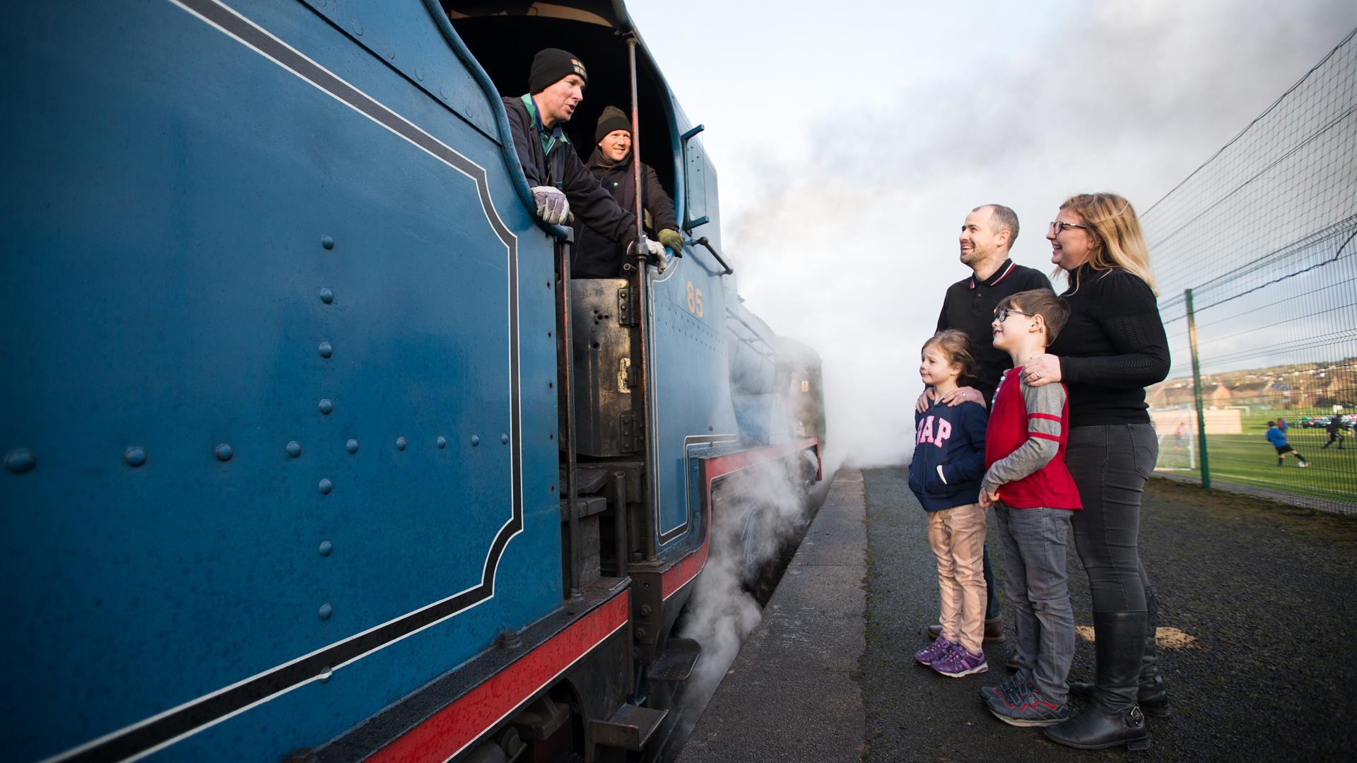 A family of four standing on the platform talking to the driver of a blue steam train