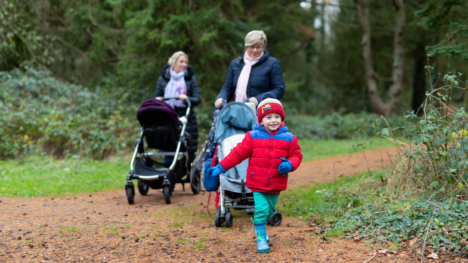 A child wearing a winter coat, hat and wellies running through the woodland. The child is followed by two adults pushing buggies.