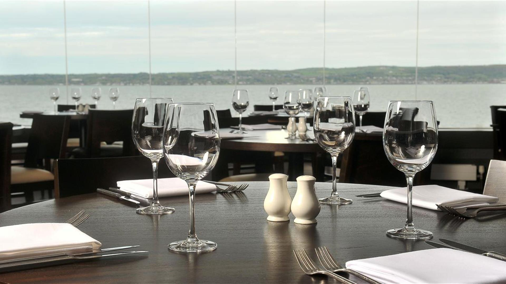 Waterfront restaurant with many tables set with wine glasses, cutlery and salt and pepper shakers.