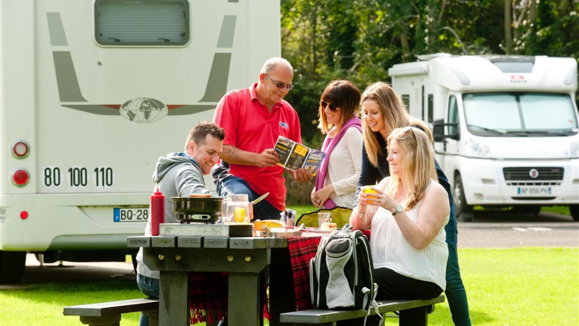 Image shows family and friends sitting at picnic table with caravan in the background