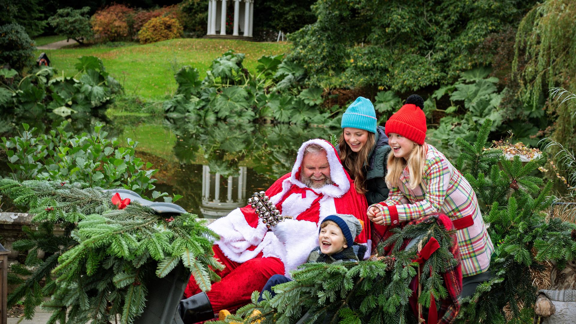 Three children are laughing and sitting with Santa in his sleigh on the grounds of Hillsborough Castle and Gardens. Santa is in a red suit.