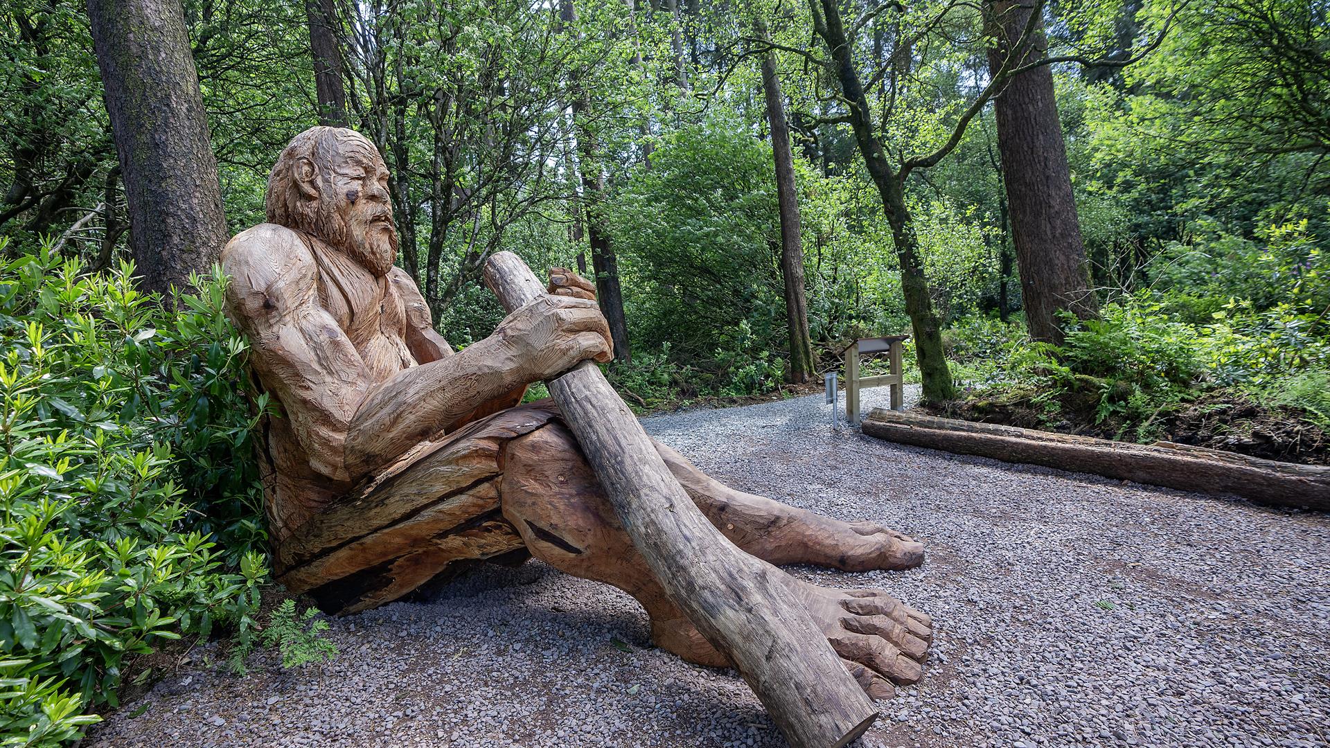 Large wooden sculpture of a giant sitting on the ground leaning against a tree holding a big stick.