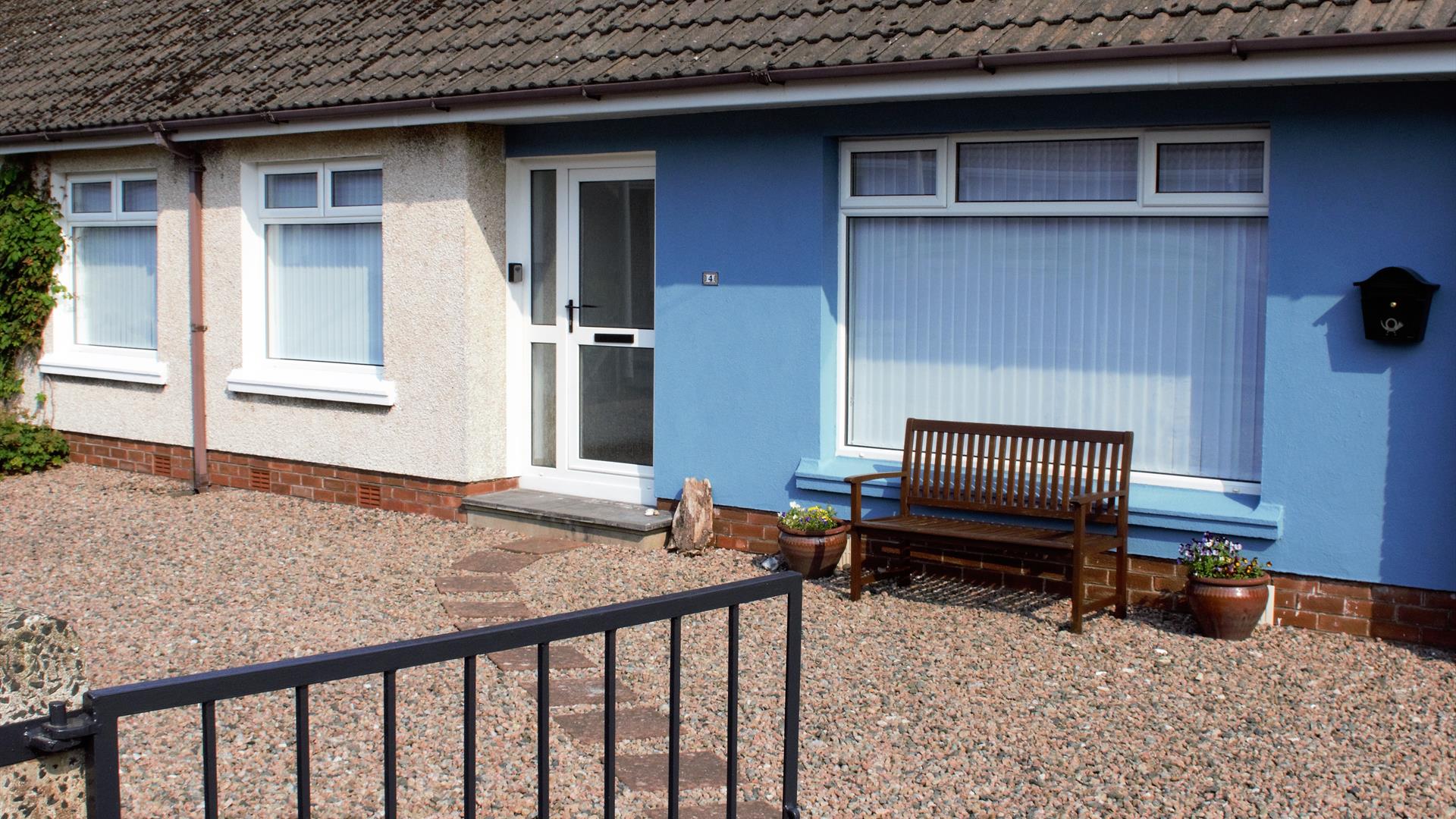 Picture depicting the external view of No.$ self-catering. A blue semi-detached house with white windows and door fronted by a gravel yard and blue ga