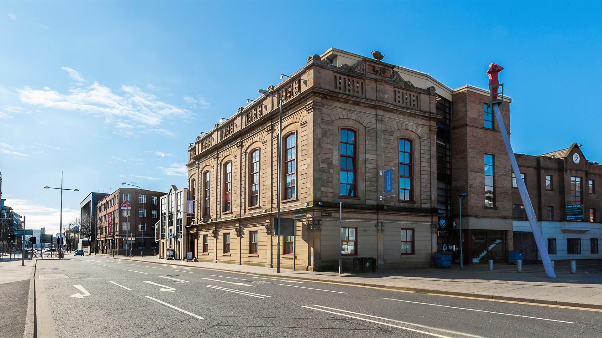 The Corn Exchange, a large light-coloured stone building with arched windows, stands on a quiet city street under a clear blue sky. There are addition