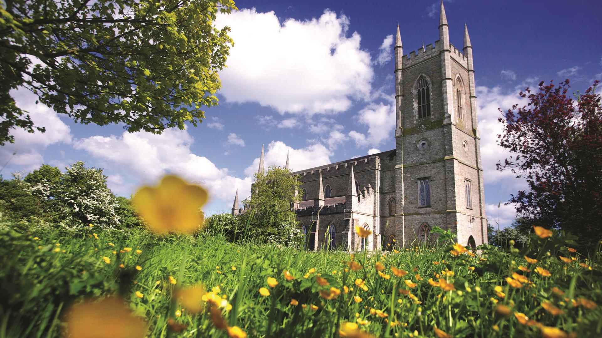 Downpatrick, Northern Ireland: The historic Down Cathedral under a blue sky, with St. Patrick's grave marker stone in the foreground, surrounded by green grass.