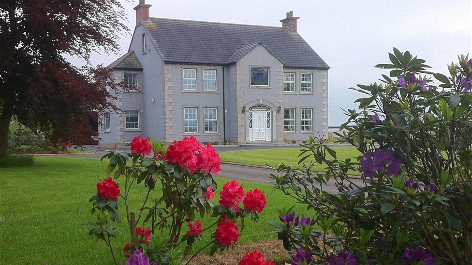 The front of a large grey house behind some colourful flowers.