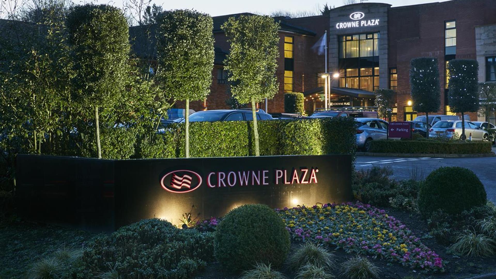 Image of the outside of the Crowne Plaza Belfast hotel. Flower bed with "Crowne Plaza" sign in the foreground, hotel and parking lot in the background