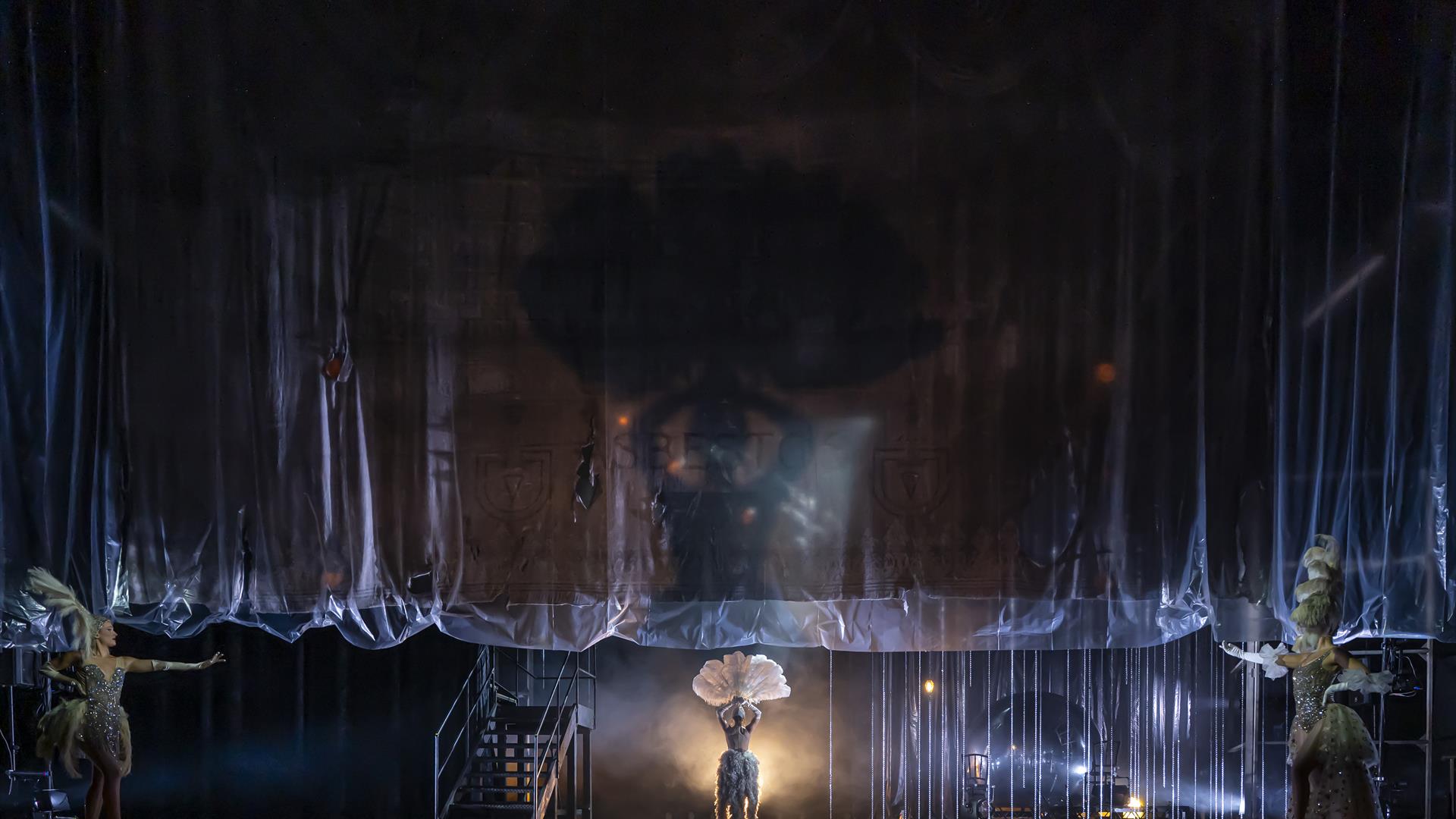 A transparent curtain rises on a dark set, with a smoky spotlight highlighting the shadow of a showgirl with a huge feather headdress walking centrall