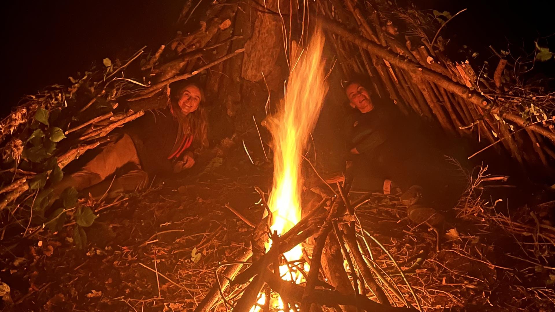 Two women laying under stick huts around a camp fire.