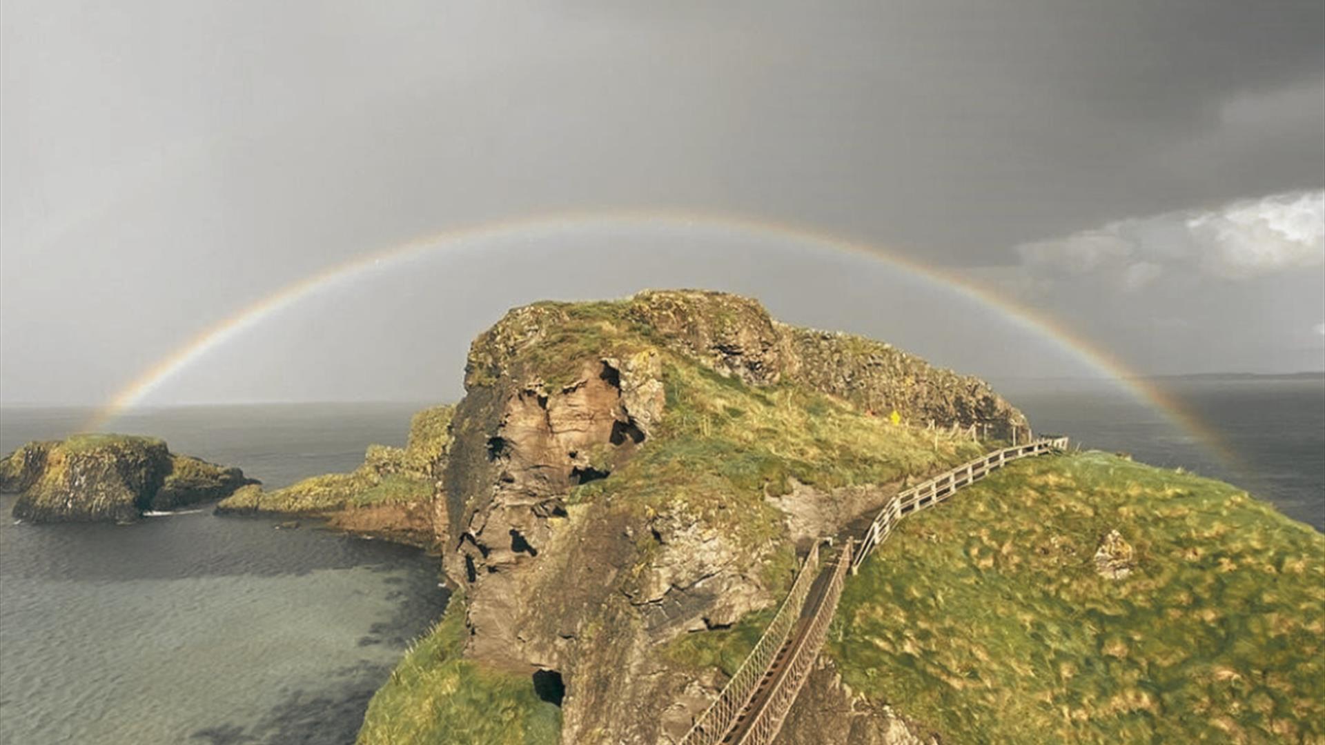 Picture of Carrick-a-Rede Rope Bridge with dark, moody grey skies and a rainbow