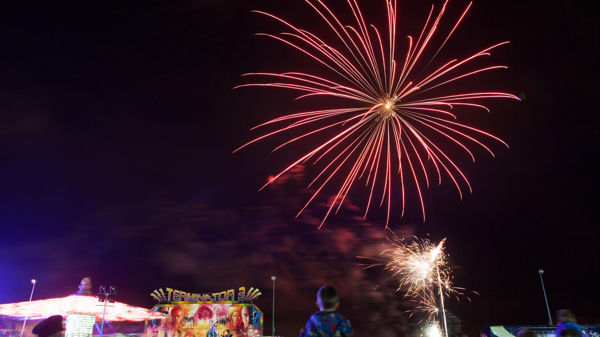 Image shows fireworks lighting up the night sky with the crowd and fairground amusements below